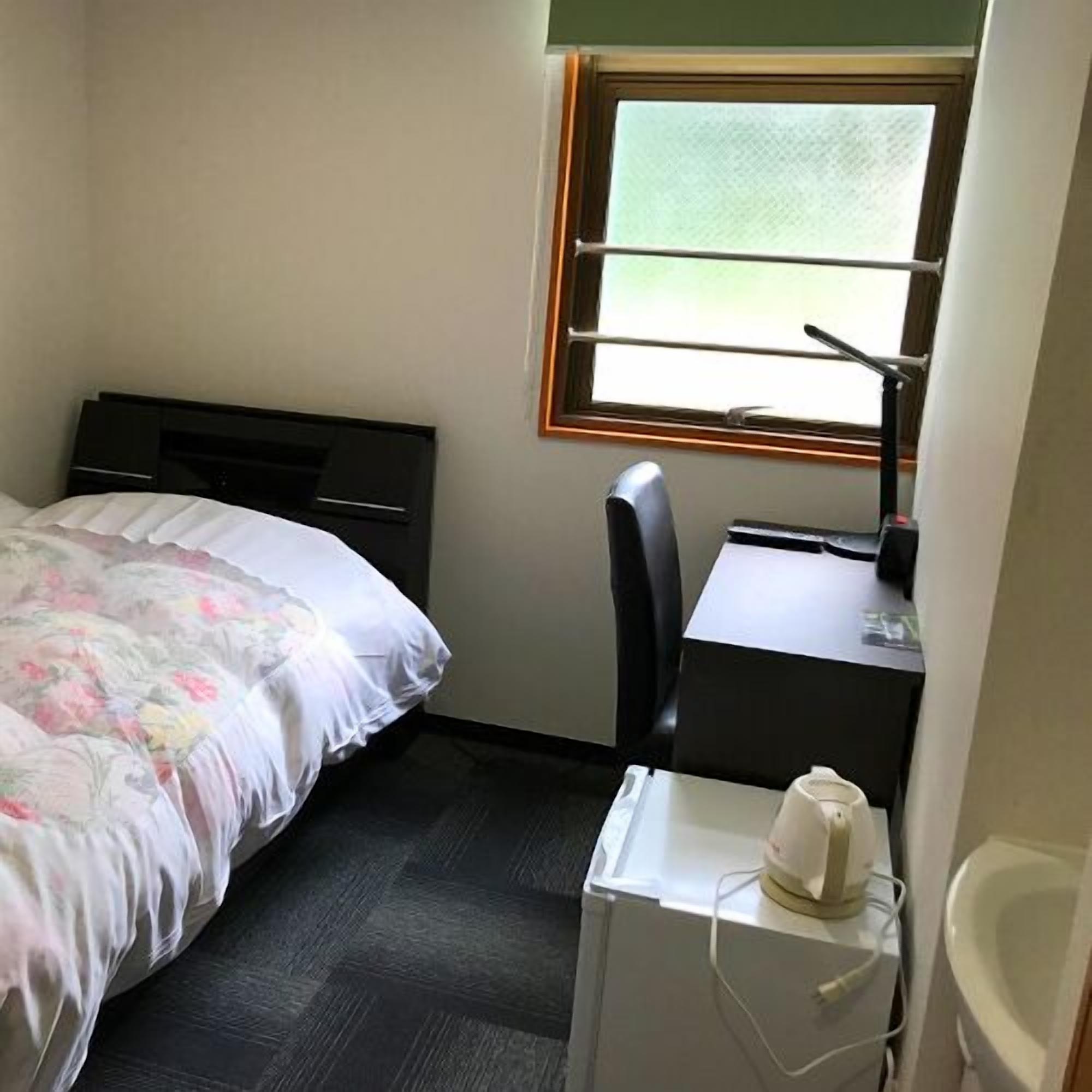 Une petite chambre d’hôtel composée d’une seule pièce, comprenant un lit, un bureau, une chaise, un radiateur et une fenêtre.