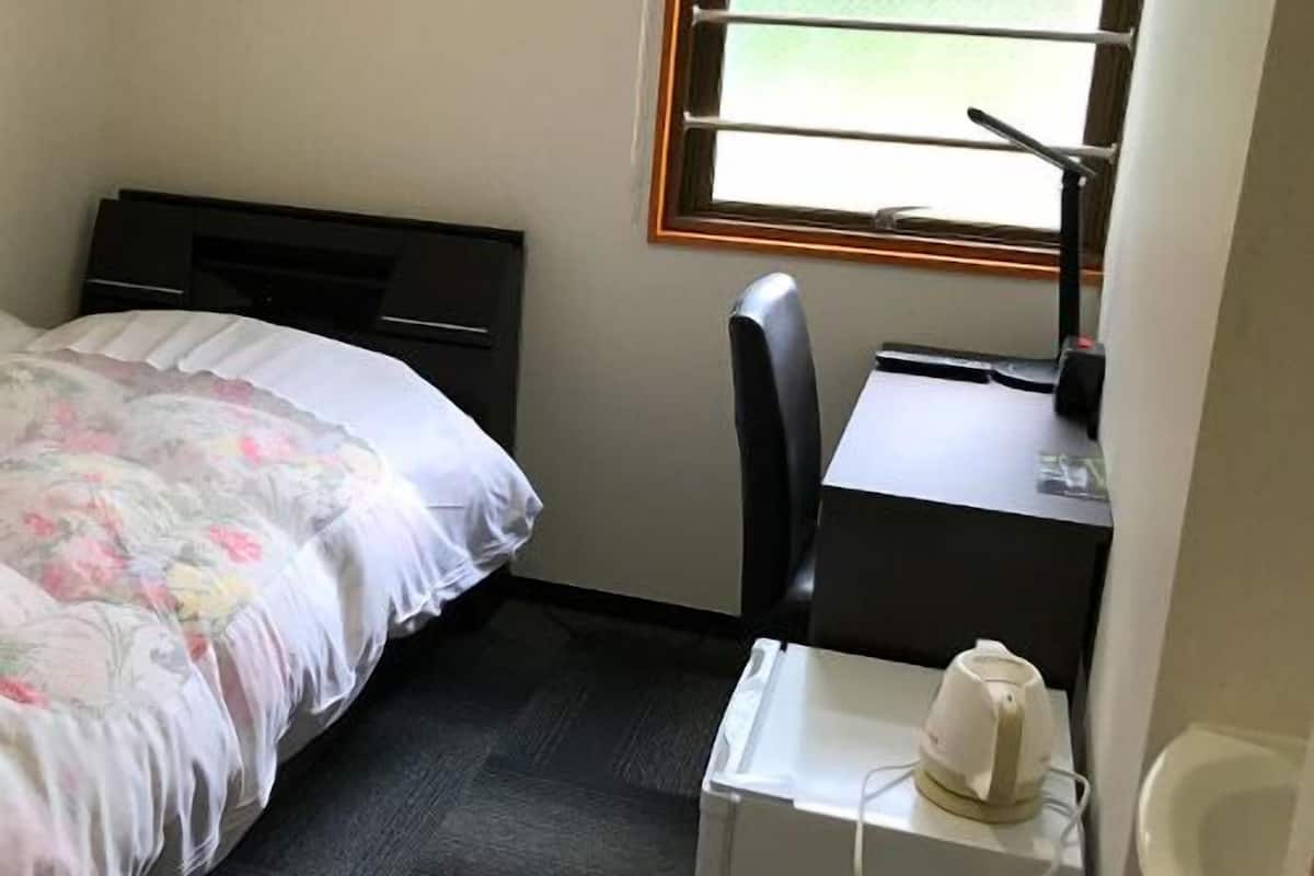 Una habitación de hotel pequeña y sencilla, con cama, escritorio, silla, radiador y ventana.