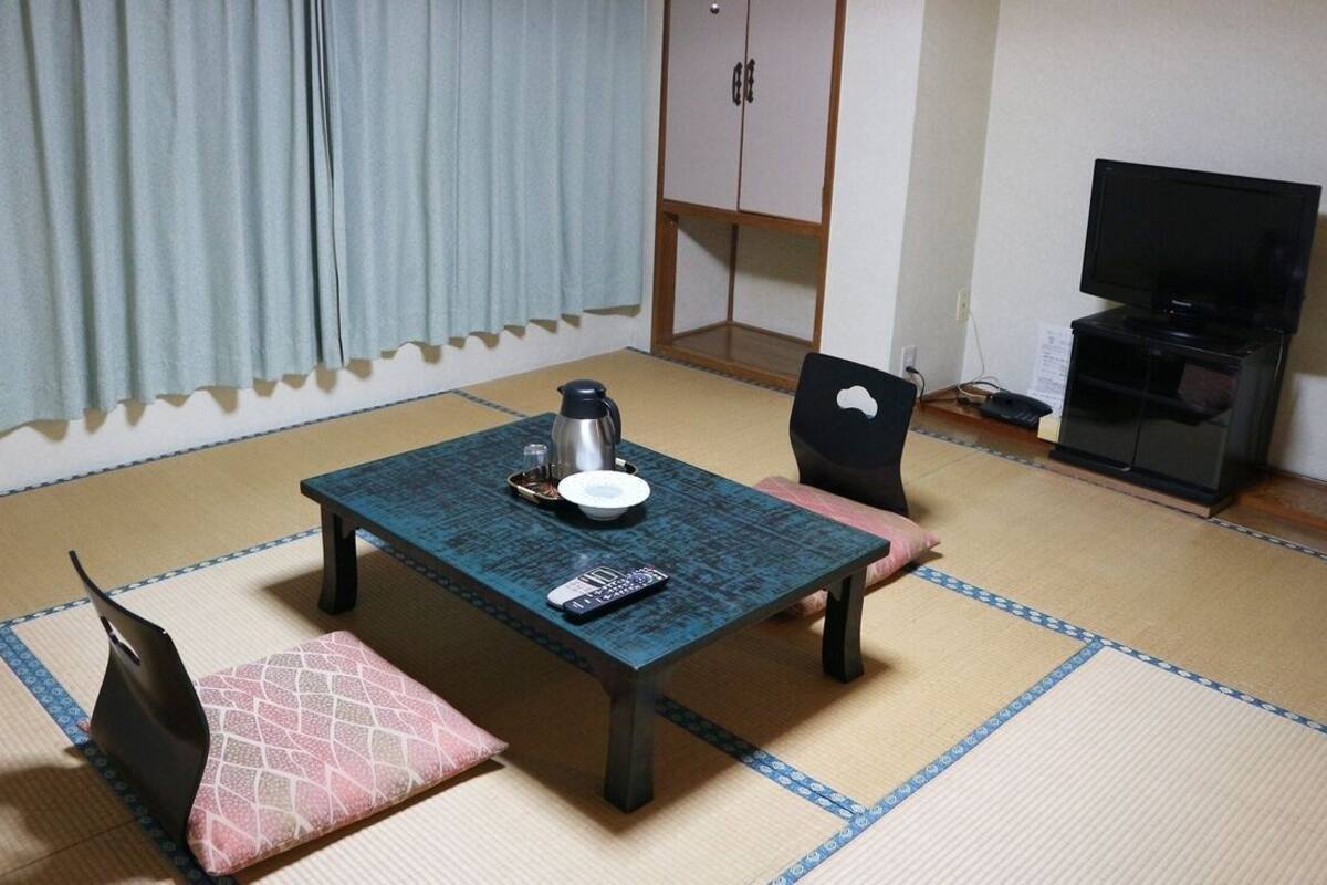 A room with tatami flooring, a table with a remote control, a kettle, and a television on a stand.
