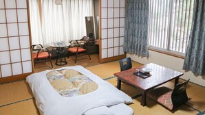 A traditional Japanese room with tatami flooring, a low table, a bed with a white mattress, and a view of the outside through a window with curtains. - Okuyama (Shinjo)