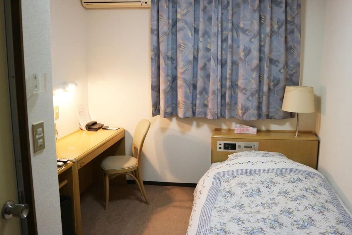 A hotel room with a bed, a desk, a chair, a lamp, and a bedside table.