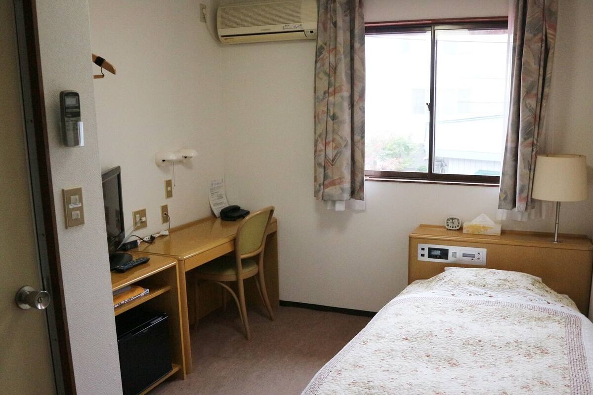 A hotel room with a bed, a desk, a chair, a desk lamp, a bedside table with a lamp, a window with curtains, and an air conditioning unit.