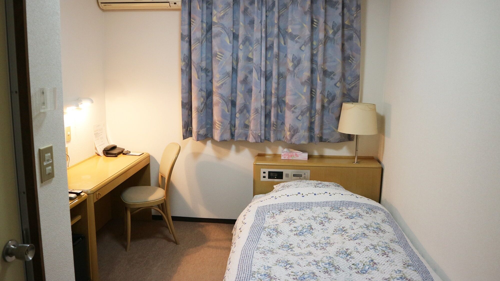 Une petite chambre d’hôtel avec un lit, un bureau, une chaise, une lampe et une fenêtre avec des rideaux.