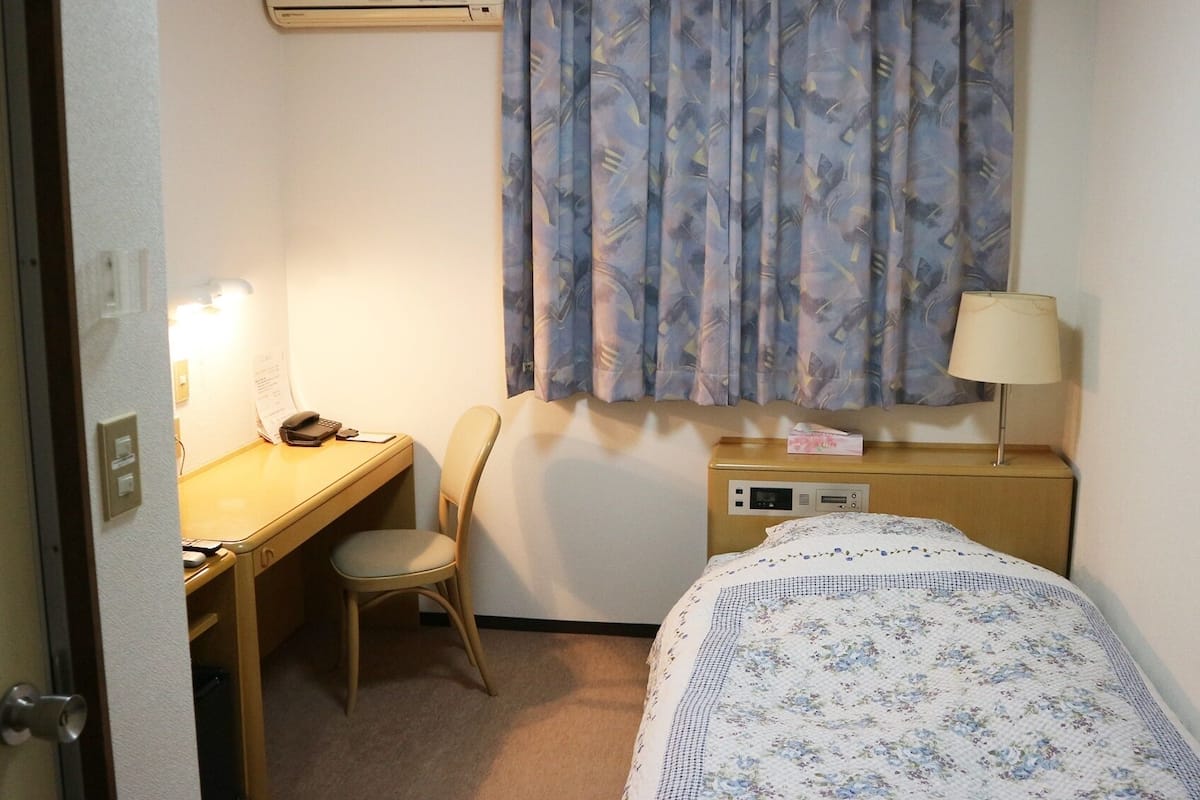 A small hotel room with a bed, a desk, a chair, a lamp, and a window with curtains.
