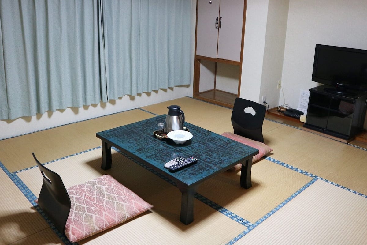 A room with tatami flooring, a small table with a kettle, a remote control, and a pink cushion.