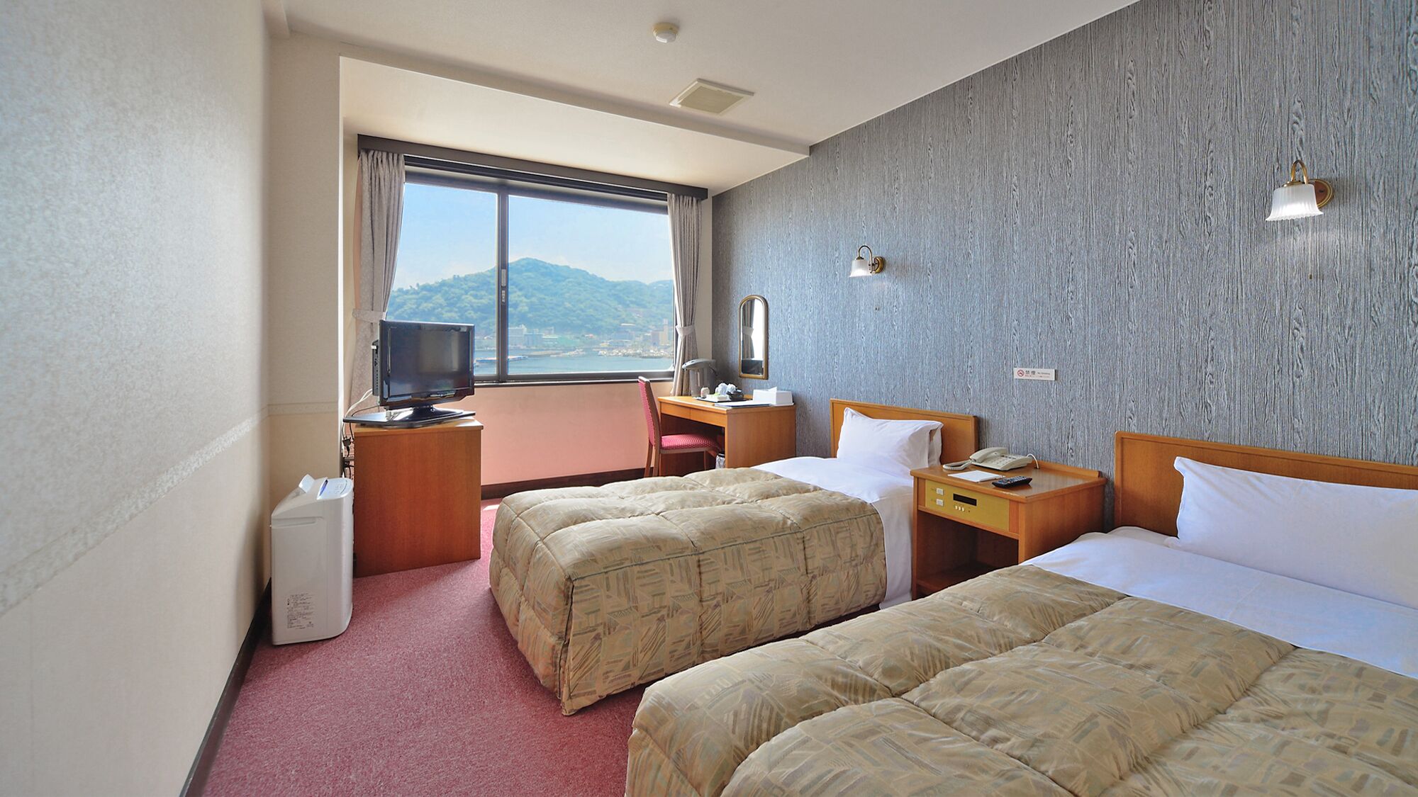 A hotel room with two beds, a desk, a television, and a view of the sea.
