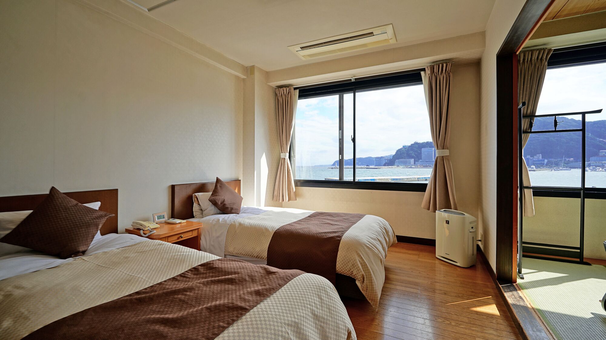 A hotel room with two beds, a window with a view of the sea, and an air conditioning unit.