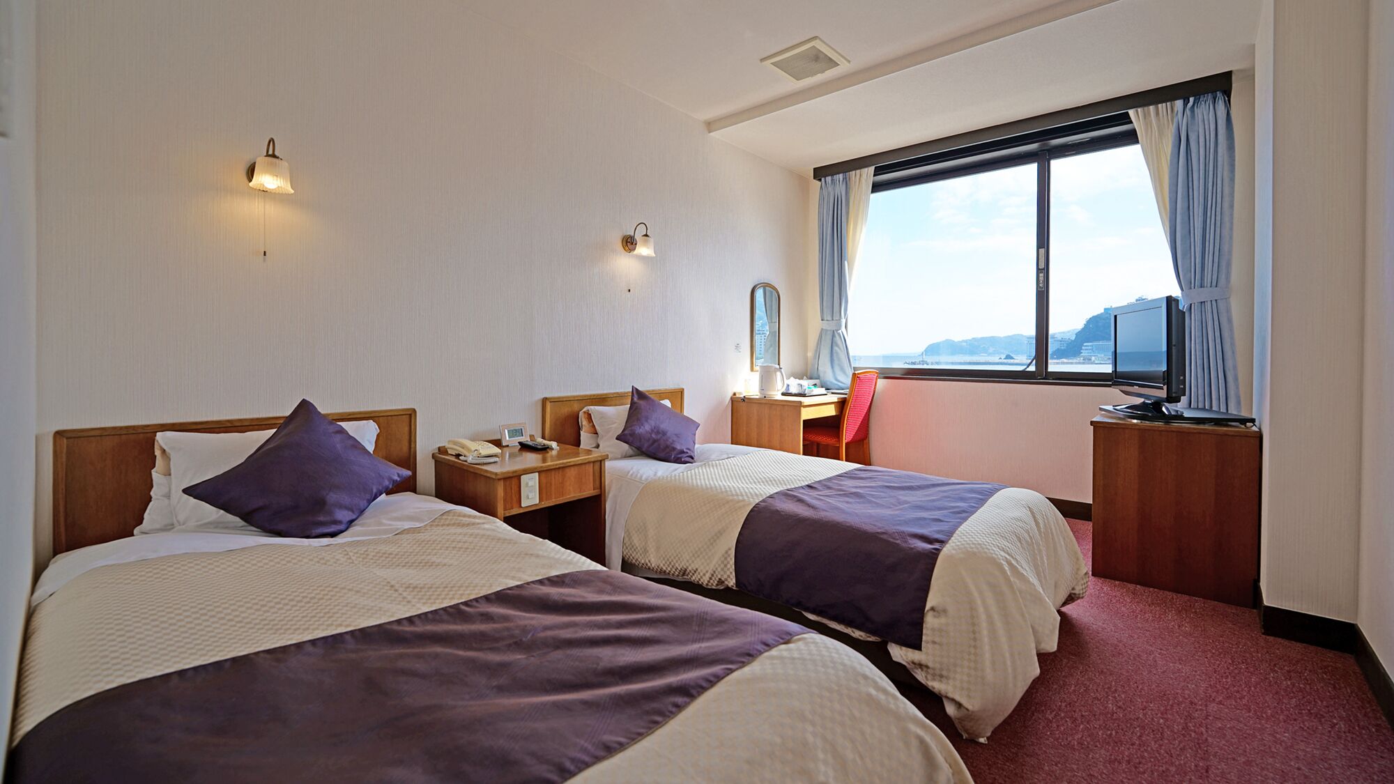 A hotel room with two beds, a desk, a chair, a TV, and a window with a view.
