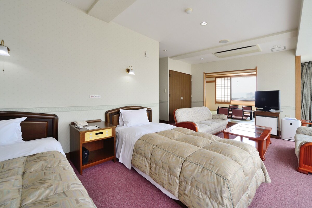 A hotel room with two beds, a desk, a chair, a TV, and a view of the city.