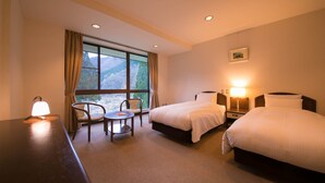 A hotel room with two beds, a desk with a lamp, a chair, and a view of mountains through the window.