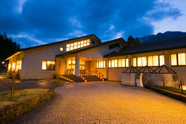 Un hotel moderno con una entrada amplia, un área exterior cubierta y vistas a la montaña al fondo.