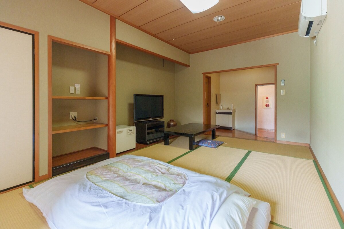 Une pièce de style japonais traditionnel, avec un sol recouvert de tatamis, un lit avec une couette, un téléviseur posé sur un meuble et une petite table.
