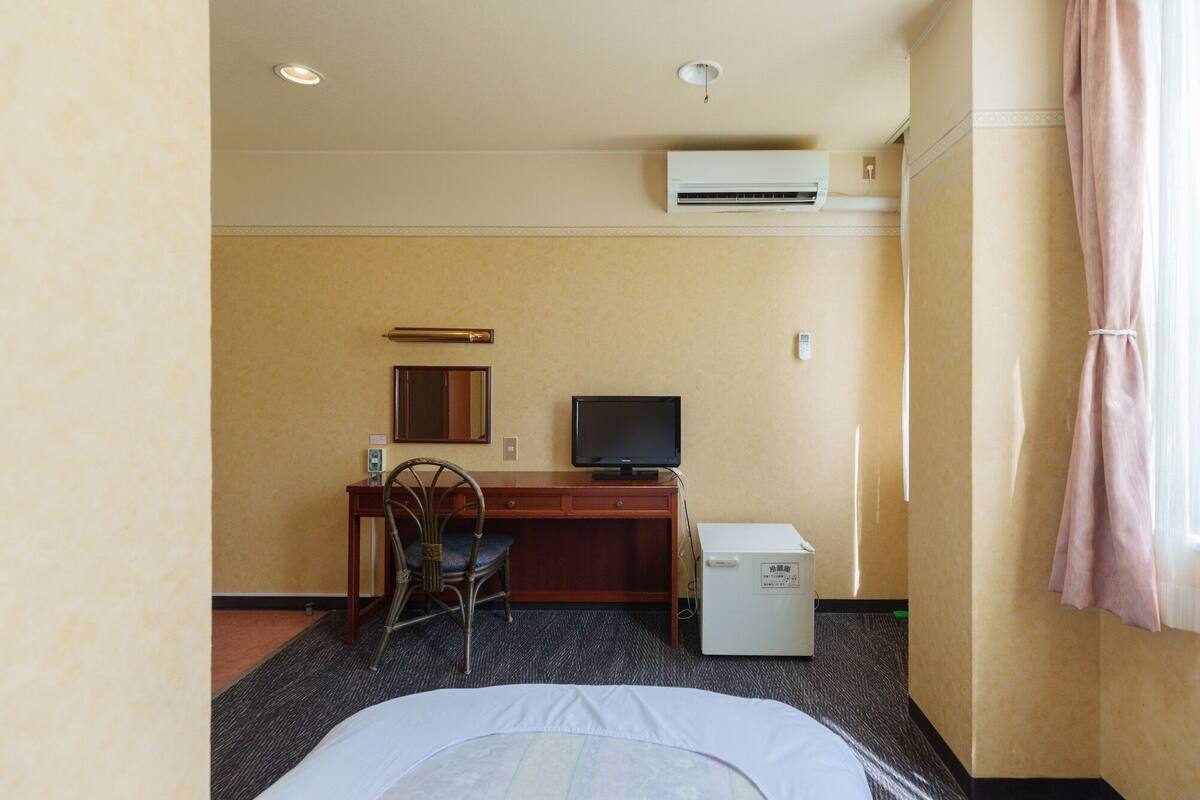A hotel room with a bed, a desk with a computer, a chair, an air conditioner, and a window with curtains.