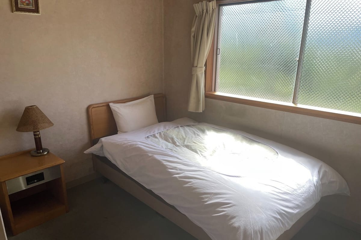 A single bed with white bedding, a wooden headboard, a bedside table with a lamp, and a window with a metal screen.