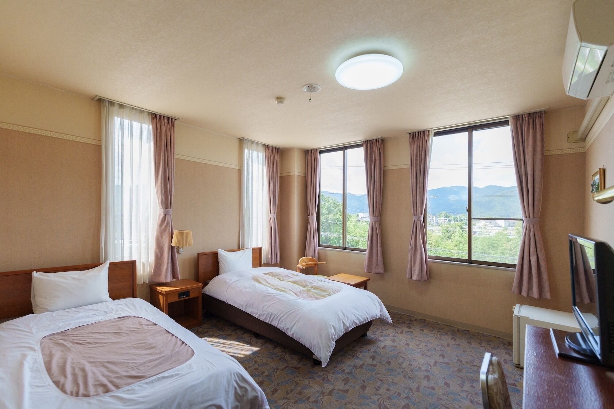 Une chambre d’hôtel avec deux lits, un bureau avec un ordinateur, une télévision et de grandes fenêtres avec des rideaux.