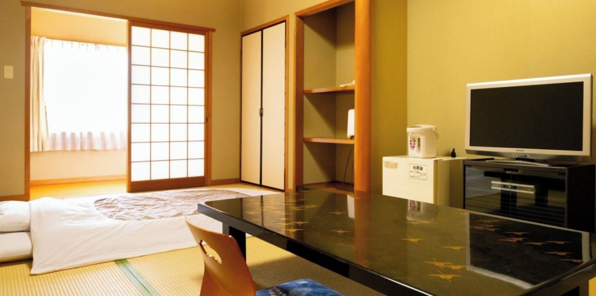 Une pièce avec un sol recouvert de tatamis, un futon servant de lit, des portes coulissantes, une télévision et un four à micro-ondes.