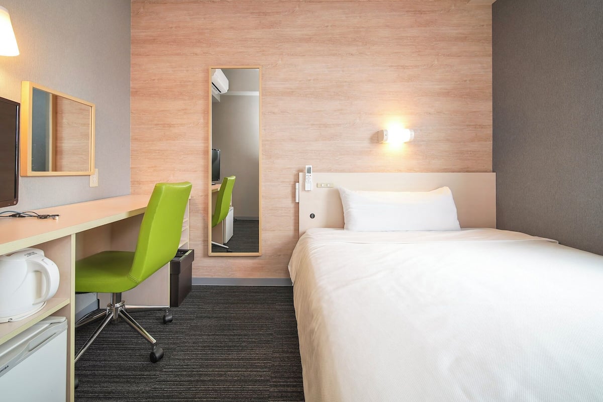 Een hotelkamer met een bed, een bureau met stoel, een spiegel, een wandlamp en een thermostaat.