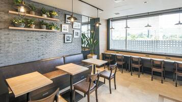 A modern cafe with wooden tables and chairs, a brick wall with plants, and large windows.