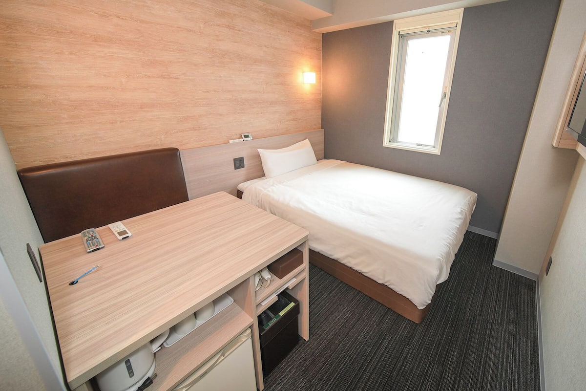 Een kleine hotelkamer met een eenpersoonsbed, een bureau met stoel, een nachtkastje, een aan de muur bevestigde televisie en een raam met zonwering.