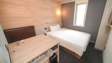 A compact hotel room with a single bed, a small desk with a chair, a bedside table, a wall-mounted TV, and a window with blinds.