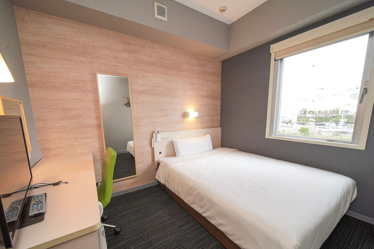 Een hotelkamer met een enkel bed, een bureau met stoel, een spiegel en een raam met uitzicht.