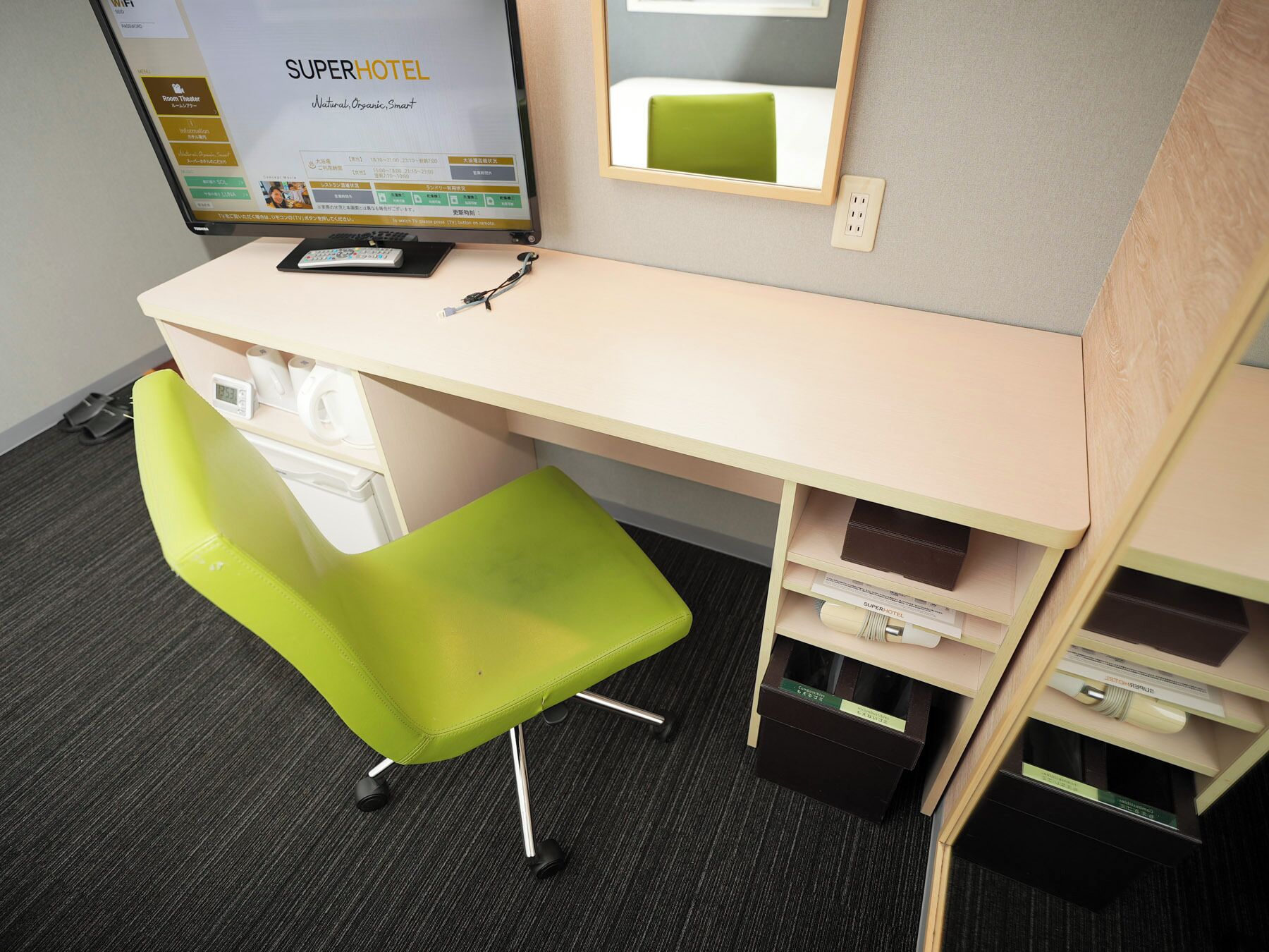 A hotel room with a desk, a bright green chair, a computer monitor displaying the SuperHotel website, a mirror, and a small shelf with a book and a remote control.