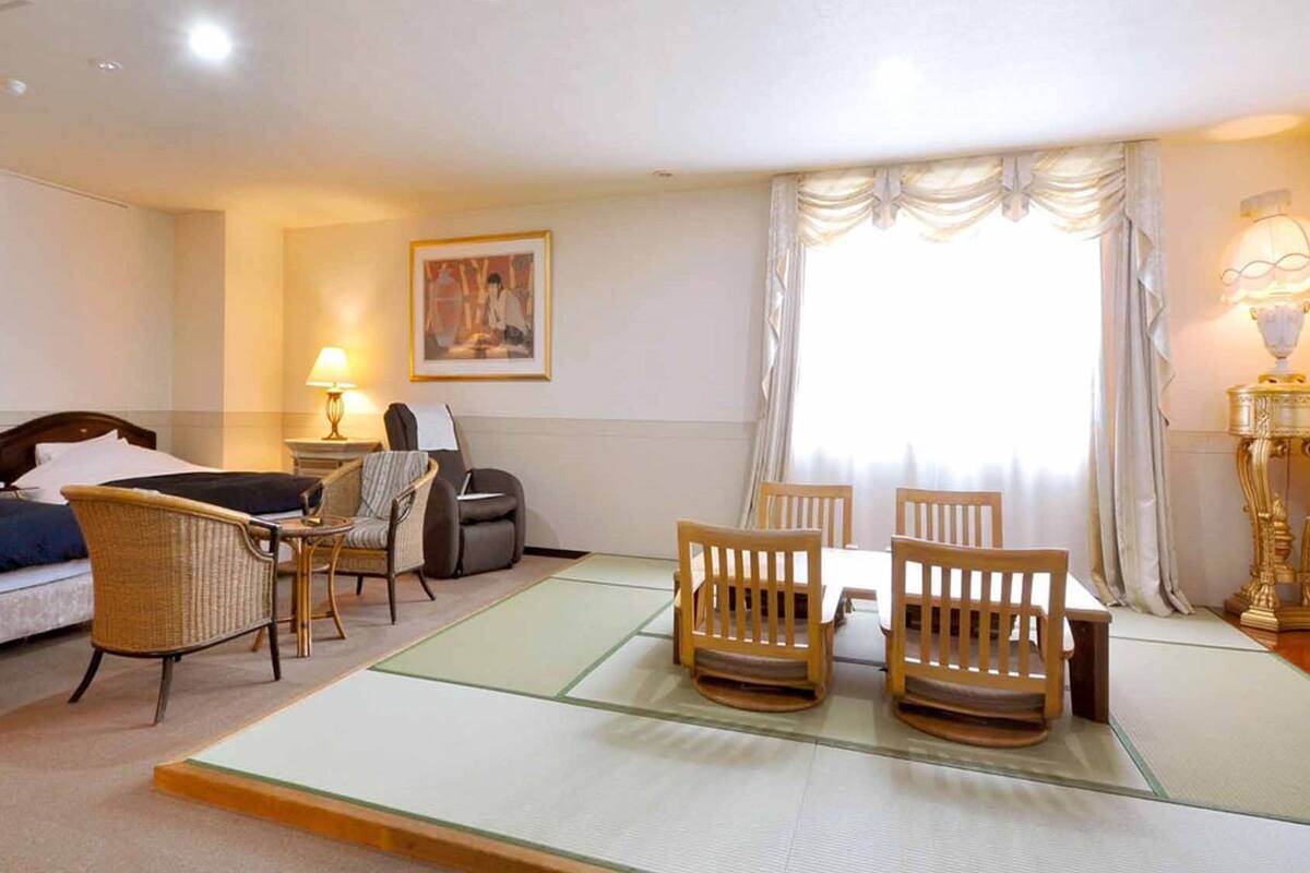 A hotel room with a bed, two chairs, a small table, a lamp, a painting on the wall, and a window with curtains.