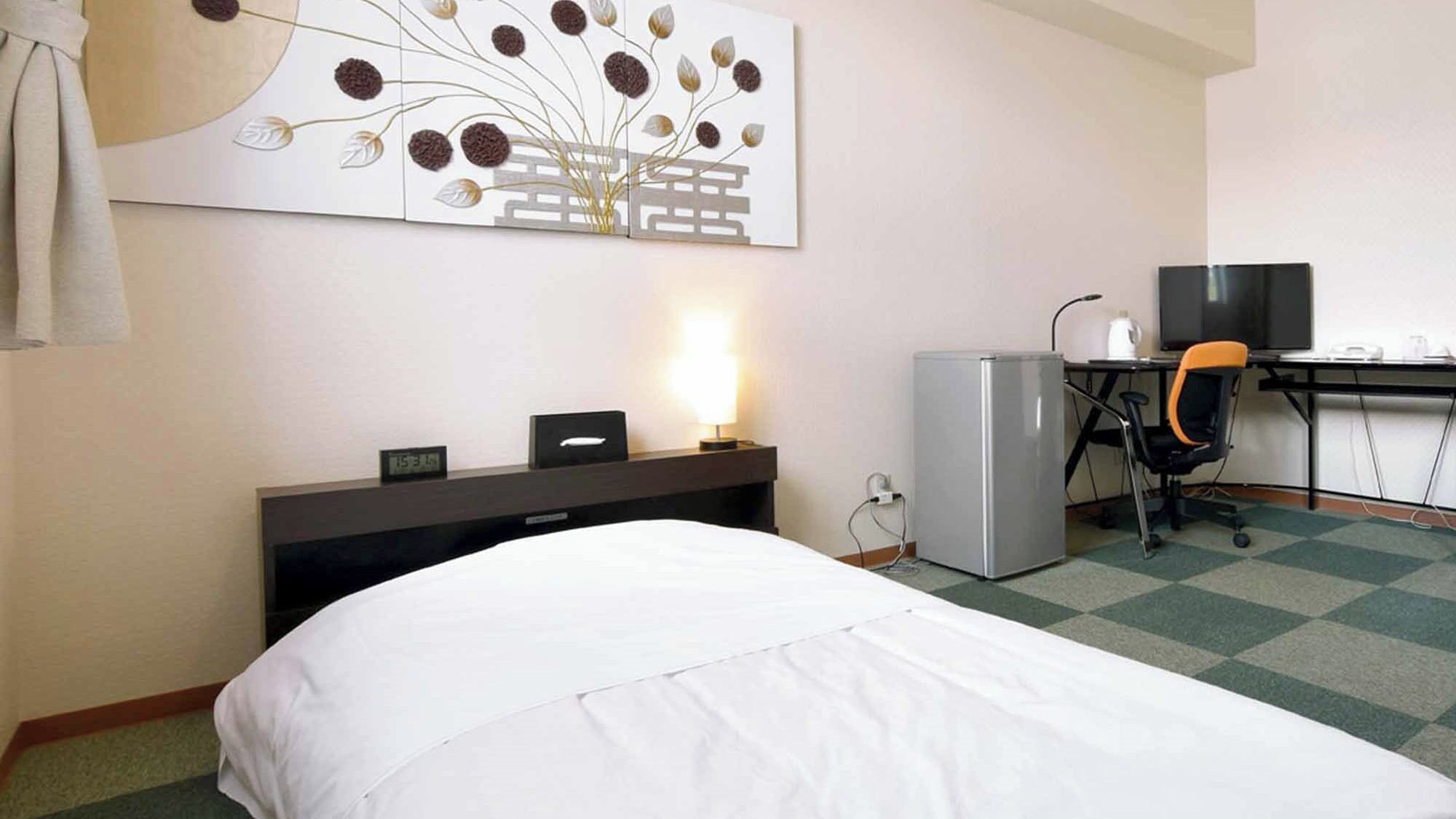 A hotel room with a bed, desk, chair, refrigerator, and a wall-mounted artwork.