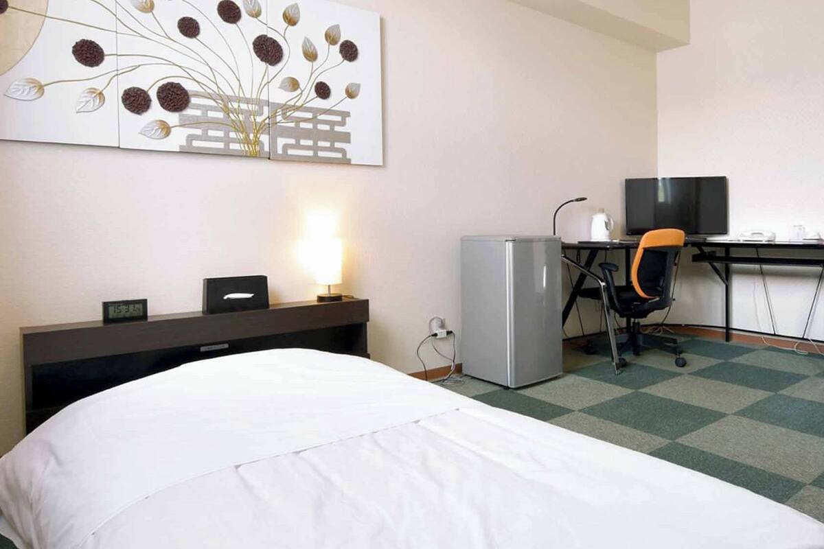 A hotel room with a bed, desk, chair, refrigerator, and a wall-mounted artwork.
