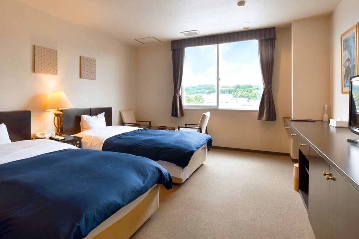 A hotel room with two beds, a desk, a chair, a window with curtains, and a TV.