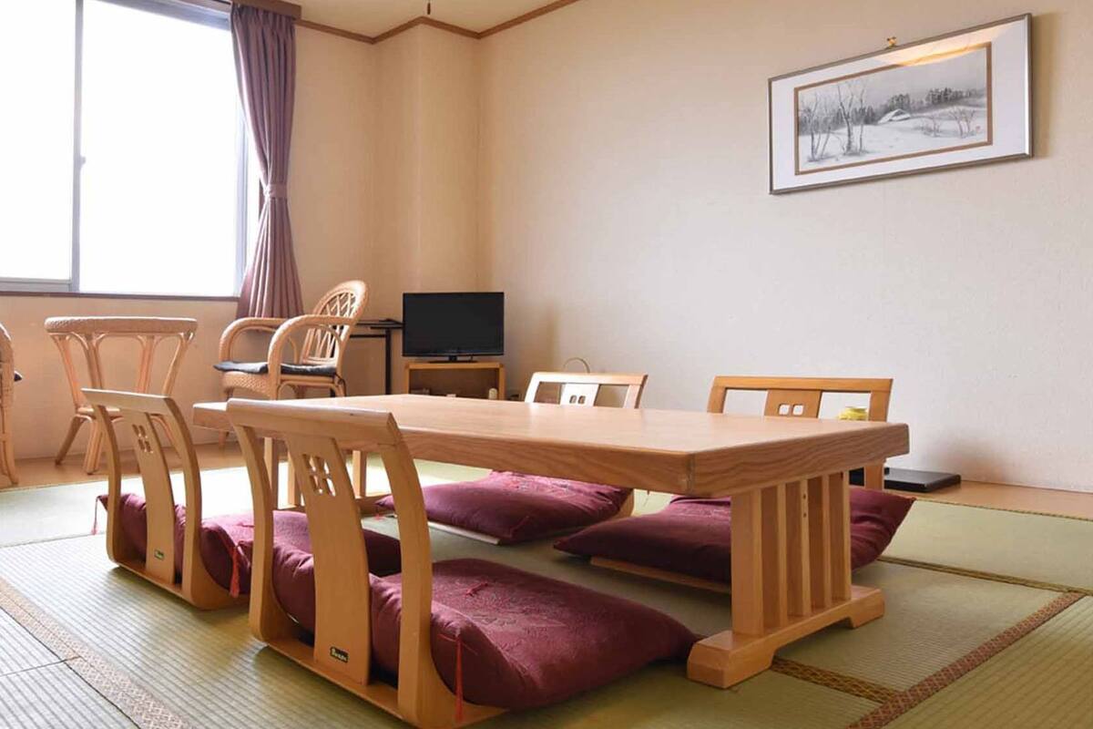 A traditional Japanese-style room with tatami flooring, a low table, wooden chairs, and a television.
