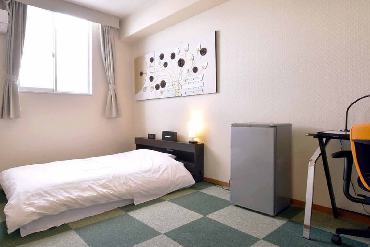 A hotel room with a bed, a desk, a chair, a refrigerator, and a wall-mounted artwork.