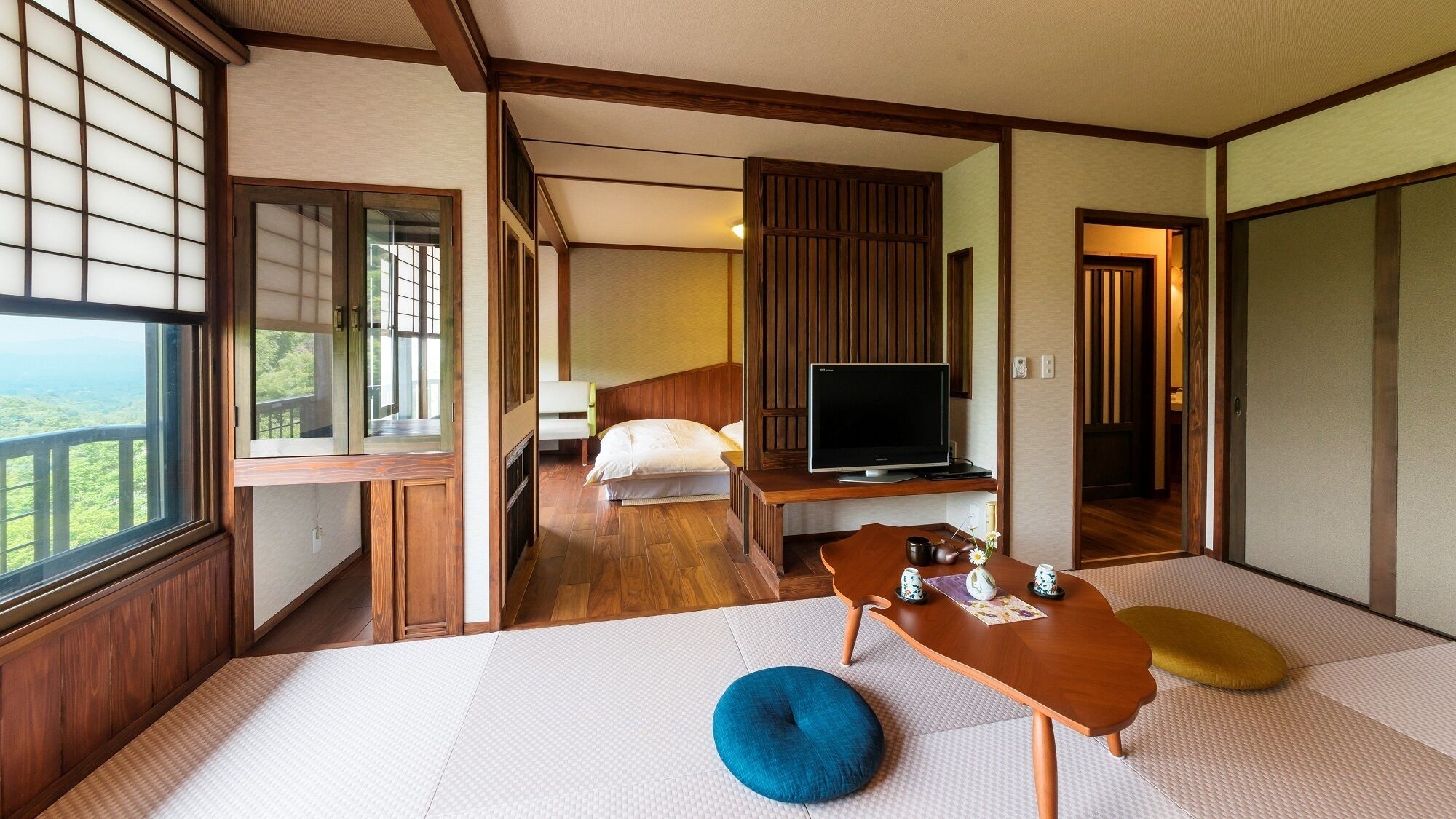 Una habitación tradicional japonesa con suelo de tatami, televisión, cama y vistas al exterior.