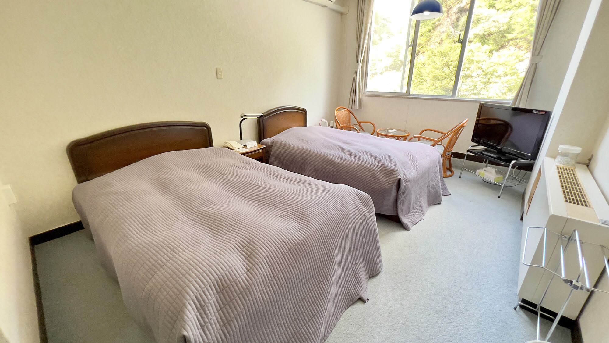 A hotel room with two beds, a desk with a computer, a chair, and a window with a view of trees.