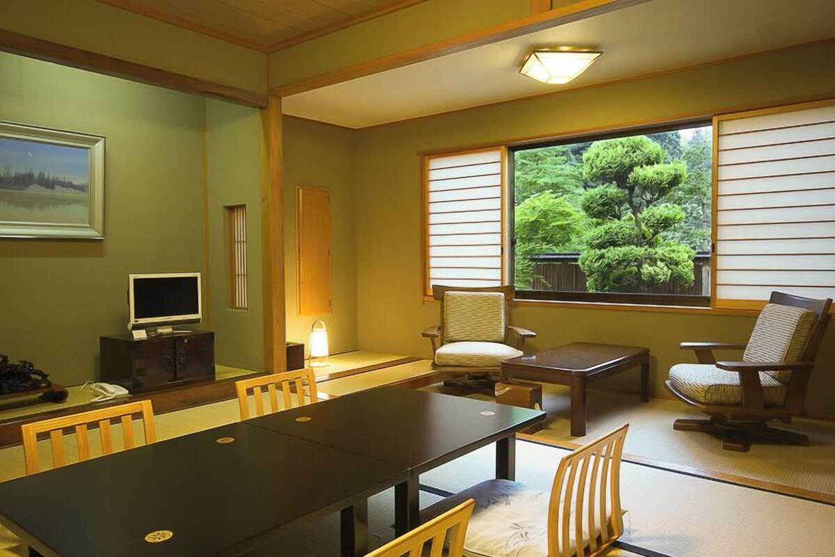 A traditional Japanese-style room with tatami flooring, a low table, chairs, a television, and a large window with a view of trees.