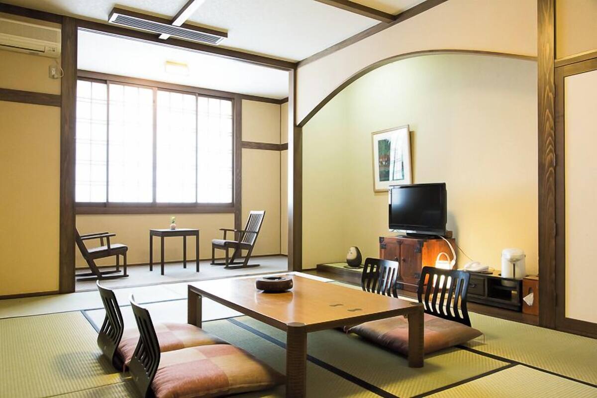 A traditional Japanese room with tatami flooring, a low table, chairs, a television, and a sliding door.