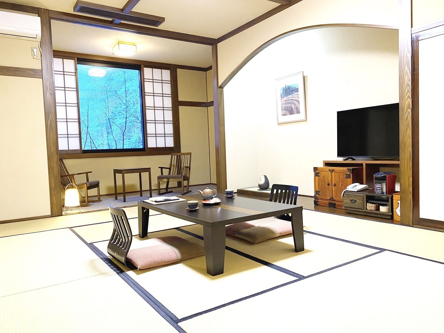 Een traditionele Japanse kamer met tatami vloerbedekking, een lage tafel, stoelen, een televisie en een raam met uitzicht op bomen.