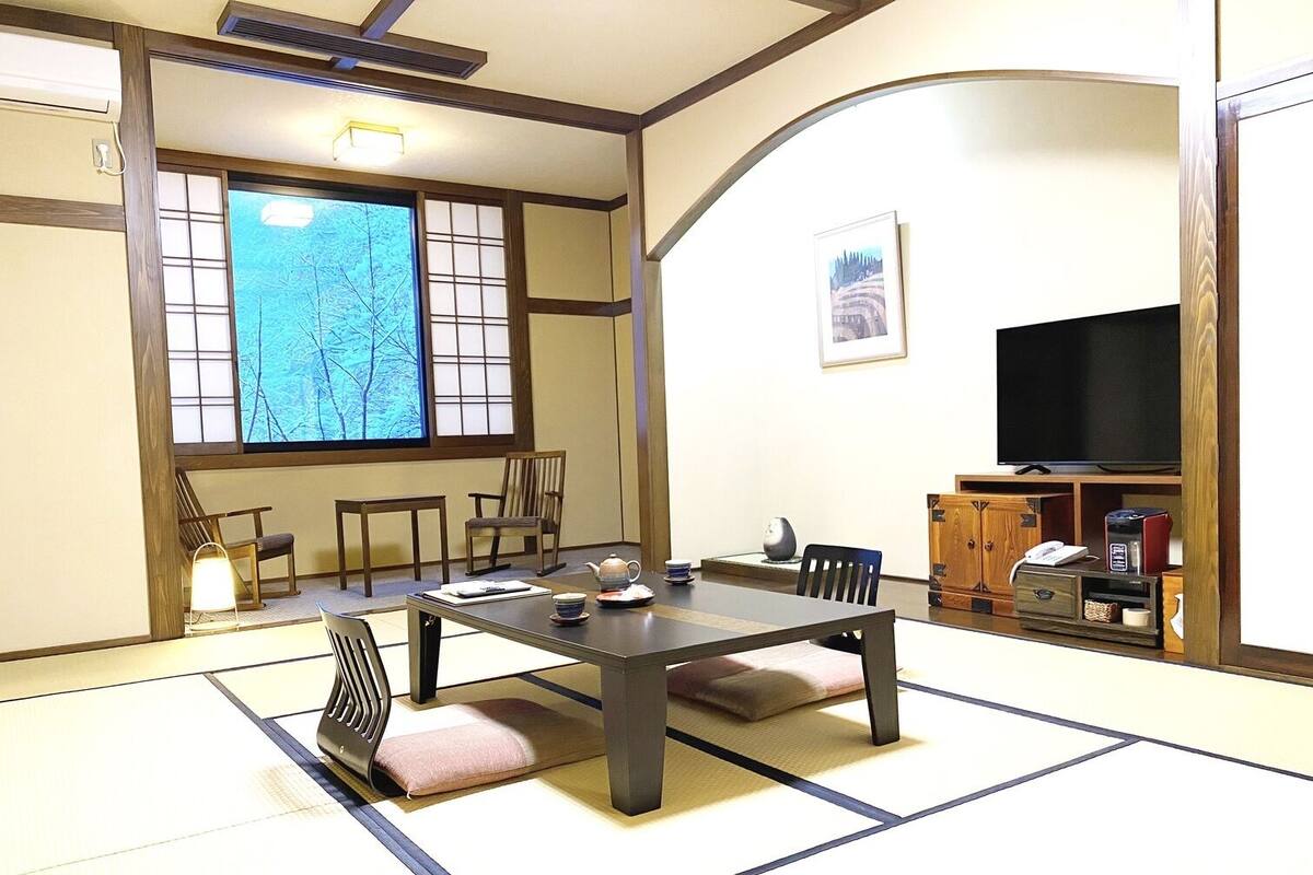 A traditional Japanese room with tatami flooring, a low table, chairs, a television, and a window with a view of trees.