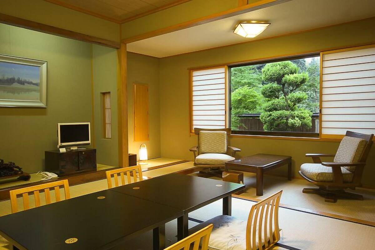 A traditional Japanese-style room with tatami flooring, a low table, chairs, a television, and a view of a lush garden.