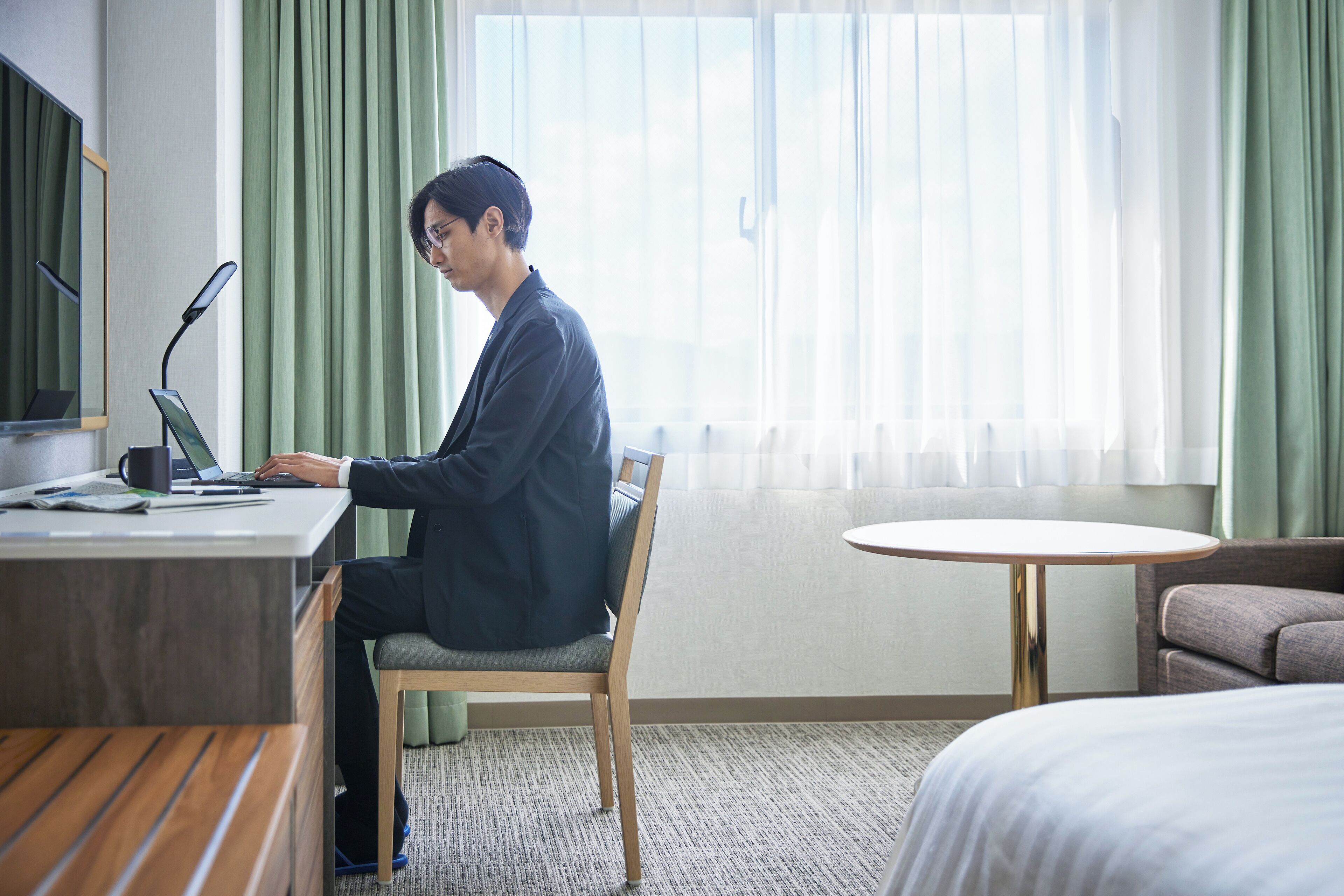 Ein Hotelzimmer mit Schreibtisch, Laptop, Stuhl, Bett und rundem Tisch.