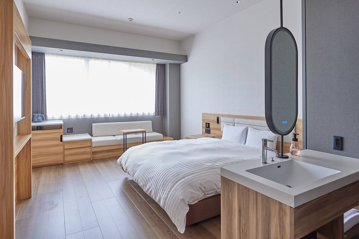 A modern hotel room with a large bed, a bedside table, a built-in desk, a large mirror, and a window with curtains.