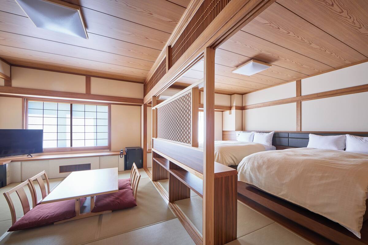 A traditional Japanese-style room with tatami flooring, a low table with cushions, a bed with a wooden headboard, and a TV on a low shelf.