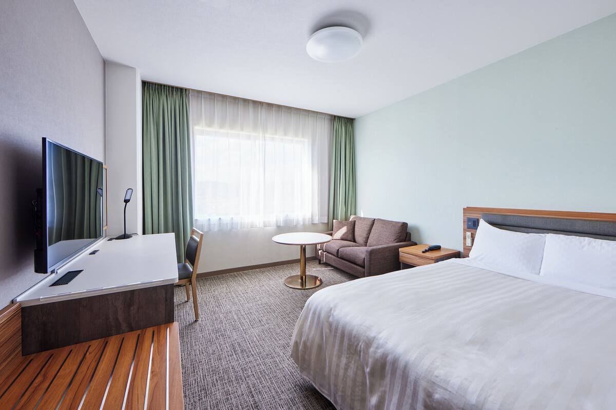 A hotel room with a bed, a desk with a computer, a TV, a sofa, and a window with curtains.