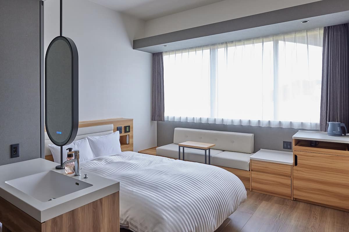 A modern hotel room with a large bed, a bedside table, a small desk, a sofa, a wooden dresser, and a large window with curtains.