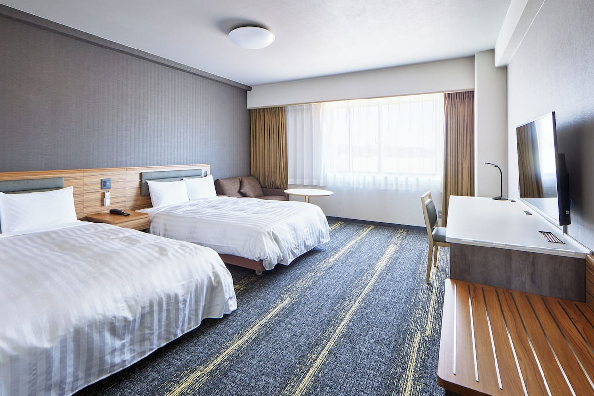 A hotel room with two beds, a desk, a TV, and a large window with curtains.