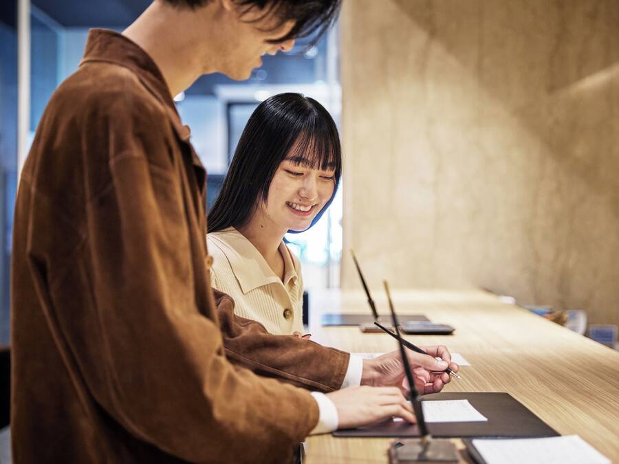 Une personne est assise à une table en bois, devant un mur bleu et sur un plancher en bois, et elle écrit avec un stylo sur une feuille de papier.