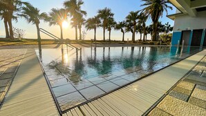 A swimming pool with a sunlit view, surrounded by palm trees and a building with a blue and white facade. - Daikoku Resort Hotel (Shibushi)