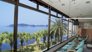 A room with large windows overlooking a body of water, a seating area with cushioned chairs, and a television mounted on the wall. - Daikoku Resort Hotel (Shibushi)