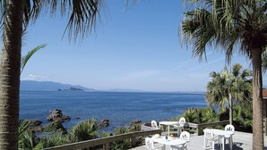 A deck with white tables and chairs overlooking a body of water with rocky outcrops and palm trees. - Daikoku Resort Hotel (Shibushi)