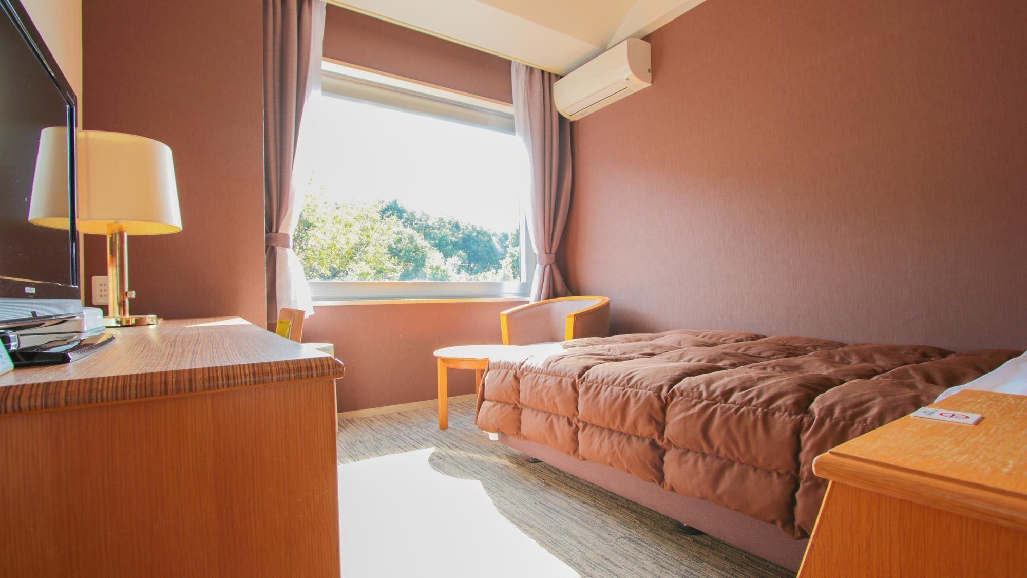 A hotel room with a bed, a desk, a chair, a lamp, and a window with a view of trees.